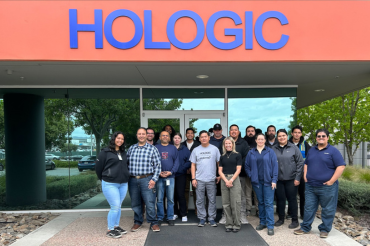 Group of people stand together in front of a sign reading "Hologic".