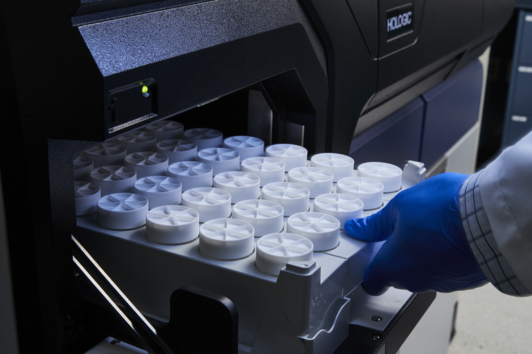 Lab technician's hand pushing a tray of small containers into a Hologic machine for testing in a lab setting.